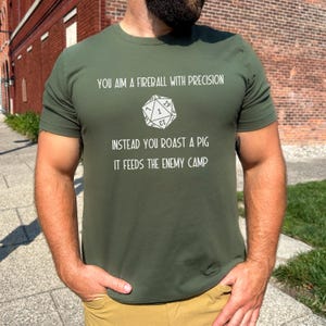 May include: Olive green t-shirt with white text and a D20 die graphic. The text reads "YOU AIM A FIREBALL WITH PRECISION, INSTEAD YOU ROAST A PIG, IT FEEDS THE ENEMY CAMP". The shirt is a short-sleeved crew neck.