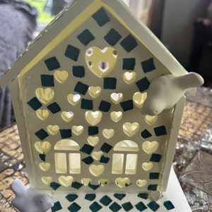 May include: A white decorative house-shaped candle holder with heart-shaped cutouts and teal mosaic accents. Two small white bird figurines are attached to the house. The house has two window cutouts and a heart-shaped cutout on the roof.