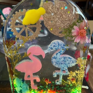 May include: A clear resin art piece featuring two flamingo figures, one pink and one blue, surrounded by floral elements, glitter, and a gold gear. A yellow shape is also embedded within the resin.