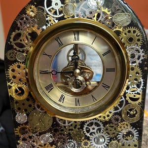 May include: A decorative clock with a gold-colored face and Roman numerals. The clock is surrounded by a black frame embedded with various gears and smaller clocks in gold and silver tones, creating a steampunk aesthetic.