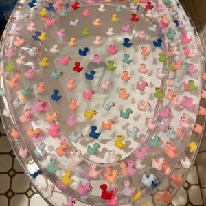 May include: A clear, oval-shaped toilet seat lid featuring a decorative inlay of numerous miniature, multi-colored rubber ducks. The ducks are arranged in a scattered pattern throughout the transparent material, showcasing various hues including pink, blue, yellow, orange, green, and red.