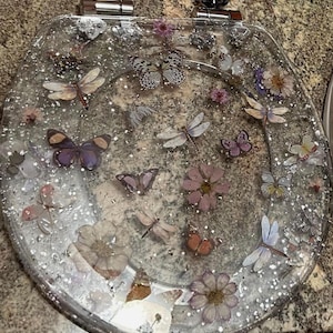May include: A clear glass toilet seat with a decorative design featuring butterflies, dragonflies, and flowers. The design is embedded in the glass and has a glittery finish.