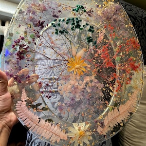 May include: A clear resin toilet seat with pressed flowers and gold leaf accents. The flowers are in shades of pink, purple, green, and yellow.