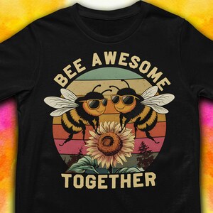 May include: Black t-shirt with a graphic of two cartoon bees wearing sunglasses, a sunflower, and the text "BEE AWESOME TOGETHER" in white. The background features a sunset design with stripes of orange, pink, green, and blue.
