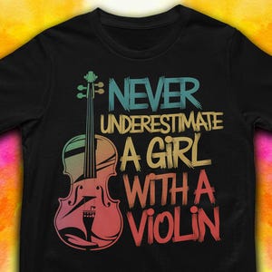 May include: Black t-shirt with a colorful graphic of a violin and the text "Never Underestimate A Girl With A Violin." The violin is depicted in shades of pink, green, and yellow. The text is in blue, yellow, and red.