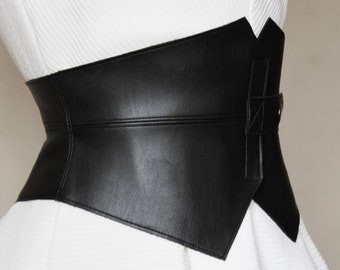May include: A wide black faux leather belt with a pointed, asymmetrical design. The belt is worn over a white dress.