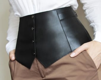 Puede incluir: Un cinturón corsé de cuero negro con un frente puntiagudo y cierres de botón. El cinturón se lleva sobre una camisa blanca y pantalones marrones.