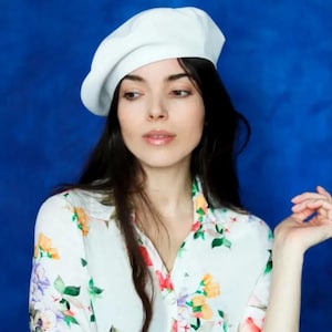 Op de afbeelding: Een vrouw met lang donker haar draagt een witte baret en een wit overhemd met bloemenprint.