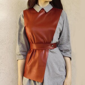May include: A rust-colored faux leather vest with a gray long-sleeved shirt underneath. The vest has a wide belt with a buckle and a pointed collar.