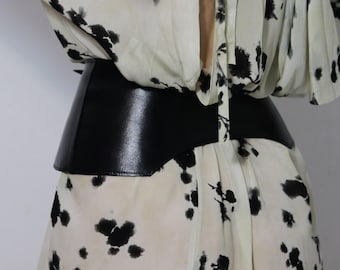 May include: A wide, black leather belt cinches the waist of a cream-colored dress with a black floral pattern. The belt has a smooth, shiny finish and a structured design, creating a defined silhouette.