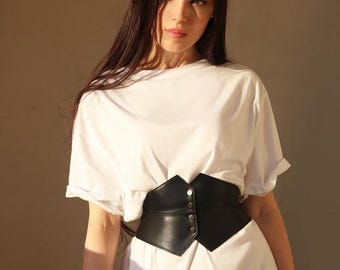 May include: A woman wearing a white t-shirt with a black leather corset belt. The belt has a pointed shape and multiple silver buckles.