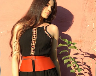 May include: A woman wearing a black sleeveless top with a geometric pattern and a wide, orange leather belt. She is standing in front of a pink wall.