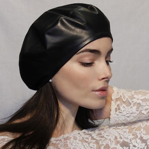Könnte beinhalten: Eine schwarze Lederbarettmütze mit glatter, glänzender Oberfläche. Die Barettmütze wird auf dem Kopf einer Person getragen, wobei ihre langen, dunklen Haare sichtbar sind.