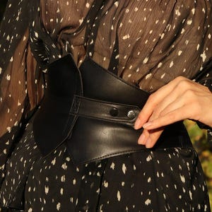 May include: A black leather corset belt with a buckle and two metal snaps. The belt is worn over a black and white patterned blouse.