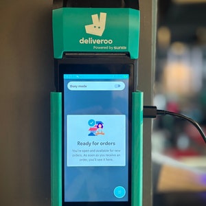 May include: A teal and black Deliveroo point-of-sale device. The screen displays "Busy mode" and "Ready for orders." The device is connected to a power source. The Deliveroo logo is visible at the top.