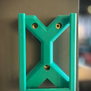 May include: A teal-coloured, geometric wall mount with a rectangular frame and a Y-shaped interior design. The mount is secured with three gold screws. The object is mounted on a grey surface.