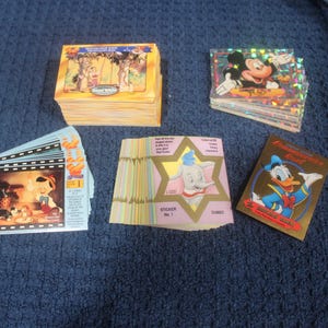 May include: A collection of Disney trading cards and stickers featuring various characters. The set includes a stack of Snow White cards, a pile of holographic Mickey Mouse cards, Pinocchio film strip cards, Dumbo star-shaped stickers, and a Donald Duck card.