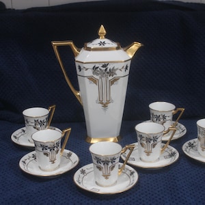 May include: A white and gold tea set with a tall, lidded teapot and six matching teacups and saucers. The set features gold trim and black floral designs. The teapot is approximately 30 cm tall.