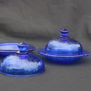 May include: Two cobalt blue glass butter dishes with matching lids. Each dish features a round base and a domed cover with a small, faceted knob handle. The glass has a textured, embossed floral pattern across the surface of the lids.