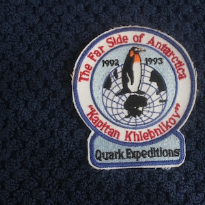Puede incluir: Parche bordado con un pingüino sobre un globo terráqueo, con texto "The Far Side of Antarctica" y "Kapitan Khlebnikov". El parche incluye los años 1992 y 1993, y "Quark Expeditions" en la parte inferior. El parche está sobre una tela azul oscuro texturizada.