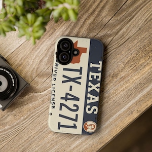 May include: A phone case featuring a Texas license plate design. The case has a cream-colored background with the state outline in red, the text "TEXAS" in white, and the license plate number "TX-4271". The case is on a wooden surface.