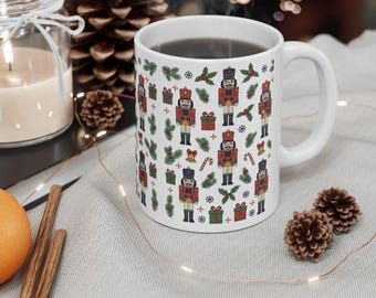 May include: A white ceramic mug featuring a repeating pattern of nutcrackers, green pine sprigs, red and green gift boxes, candy canes, golden bells, and small blue snowflakes. The mug has a standard handle and is filled with a dark liquid.