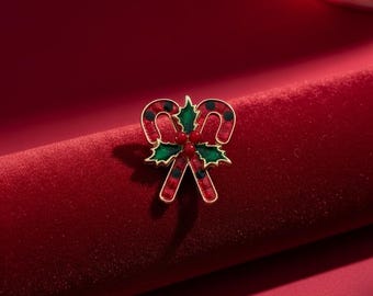 May include: A gold-toned Christmas brooch in the shape of a candy cane and holly leaves. The candy cane is red with black dots, and the holly leaves are green. The brooch is set against a red velvet background.