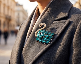 May include: A swan-shaped brooch featuring a gold-tone metal frame encrusted with clear white crystals along the neck and head, with a body composed of multiple marquise-cut emerald green gemstones pinned to a dark gray wool coat.