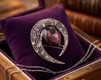 May include: A silver-toned brooch with a large, teardrop-shaped, deep purple gemstone at its center. The brooch is set on a purple velvet cushion inside a wooden jewelry box, with a silver chain visible.