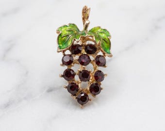 May include: A gold-toned grape cluster pendant with a green enameled leaf design. The pendant features a cluster of dark red gemstones, likely garnets, set in a gold metal frame. The leaves are a bright green color.