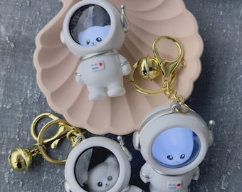May include: White astronaut keychains with gold-colored accents. Each keychain features a small, cartoon-like astronaut figure with a clear visor, revealing a small animal inside. The keychains are on a pink seashell-shaped dish.