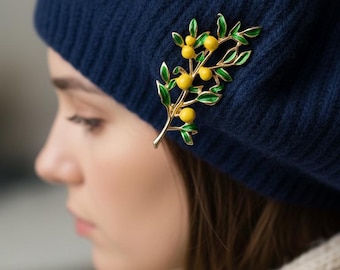May include: A navy blue beret adorned with a gold-toned brooch featuring a branch with yellow fruit and green leaves. The brooch adds a touch of elegance to the hat. The beret is made of a soft, textured material.