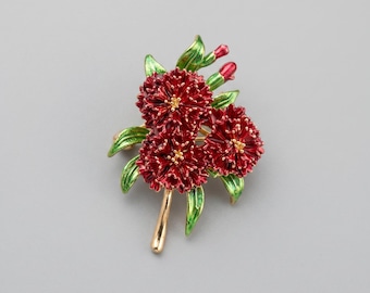 May include: A floral brooch featuring three red flowers with gold centers and a gold stem. The brooch has green leaves and red buds. The brooch is a decorative accessory.