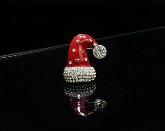 May include: A festive red Santa hat brooch with a sparkling crystal-studded base and a large, clear crystal at the tip. The hat is adorned with gold stars and small crystals, perfect for holiday wear.