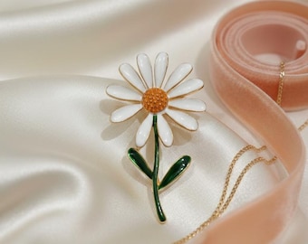 May include: A daisy flower brooch with white petals, a gold-colored rim, and an orange center. The stem and leaves are green. A gold necklace and a pink velvet ribbon are also in the image, set against a white satin background. The brooch is a jewelry item.