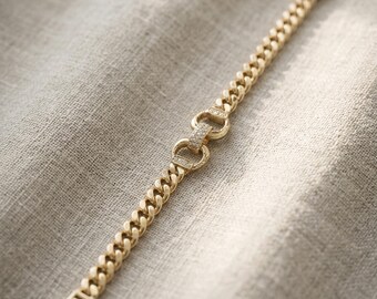 May include: A gold-colored bracelet with a curb chain design. The bracelet features a clasp closure and a decorative center piece with small, sparkling embellishments. The jewelry is displayed on a neutral-toned fabric background.