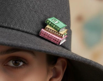 May include: A gray fedora hat with a stack of three jeweled book pins. The pins are in shades of pink, yellow, and green, with sparkling details. The hat is worn at an angle, with the wearer's eye and ear visible.