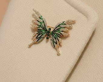 May include: A gold-toned butterfly brooch with green and white accents. The butterfly's wings are detailed with green enamel and sparkling white crystals. The brooch is pinned to a cream-colored fabric.