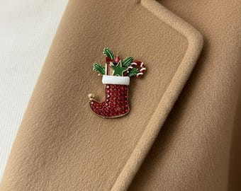 May include: A Christmas stocking brooch with red rhinestones, a white cuff, and gold trim. The stocking is filled with a green star, candy canes, and sprigs of evergreen. The brooch is pinned to a tan blazer.