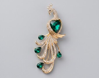 May include: Gold-toned peacock brooch with a large emerald green heart-shaped gemstone and smaller teardrop-shaped green gems. The peacock's body and tail feathers are adorned with clear crystals.