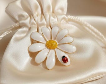 May include: A white daisy flower brooch with a gold outline and a yellow center, adorned with a small red ladybug. The brooch rests on a cream-colored satin drawstring bag, creating a delicate and elegant aesthetic.