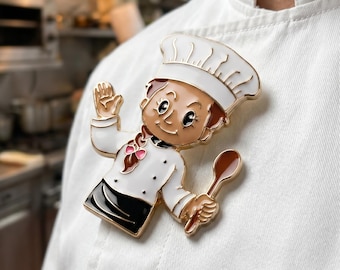 May include: A colorful chef pin featuring a cartoon chef with a white hat and uniform, holding a spoon. The pin is gold-toned with black, white, brown, and pink accents. The pin is attached to a white chef's coat.