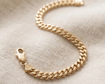 May include: Gold-colored curb chain bracelet with a lobster clasp. The bracelet is made of interlocking oval links. The clasp has the initials "XP" engraved on it. The bracelet is displayed on a neutral-colored fabric.