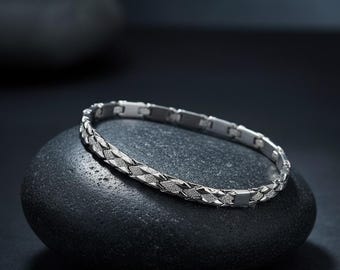 May include: A silver bracelet with a geometric pattern rests on a black stone. The bracelet features a series of rectangular and diamond-shaped links, creating a textured appearance. The jewelry is displayed against a dark background, highlighting its metallic shine.