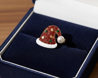 May include: A festive Santa hat brooch, featuring red and green gemstones, gold stars, and a pearl accent. The hat is set in a gold-tone metal and rests in a navy blue jewelry box. A charming holiday accessory.
