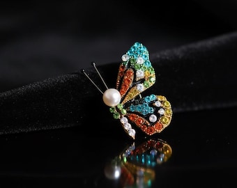 May include: A colorful butterfly brooch with a pearl body and wings adorned with vibrant gemstones. The wings display a gradient of colors, including blue, green, yellow, and orange. The brooch is set against a black background, creating a striking contrast.