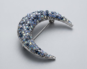 May include: A crescent moon brooch with a silver-tone metal base, encrusted with blue and iridescent crystals. The brooch has a pin on the back for secure fastening. This celestial-themed jewelry piece is suitable for various occasions.