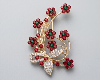 May include: A gold-toned brooch featuring a floral design with red and green gemstone accents. The brooch has a bow and leaf design with clear crystal embellishments. The flowers have red petals and green centers.