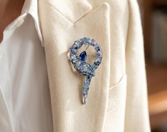 May include: A bejeweled brooch in the shape of a snake, featuring a deep blue teardrop-shaped stone at its center. The snake's body is adorned with a variety of blue and purple stones. The brooch is pinned to a cream-colored blazer.