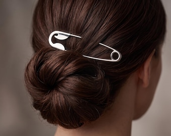 May include: A large, silver-toned metal hair accessory shaped like a safety pin. The pin is positioned horizontally, securing a dark brown hair bun. The accessory features a classic safety pin design with a pointed end and a coiled spring base.